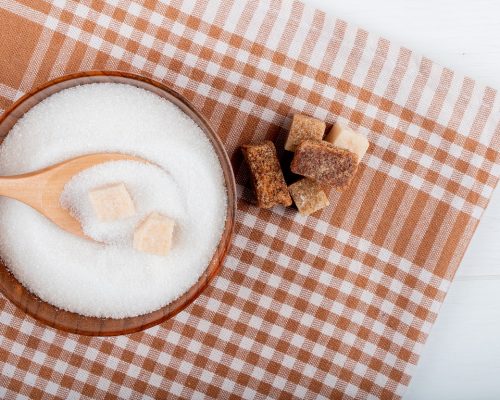 top view of white sugar in a wooden bowl with a spoon and lump sugar and palm sugar pieces on plaid tablecloth with copy space